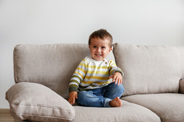 Portrait of adorable little boy sitting on the textile couch and smiling. Happy toddler laughing and being playful at home. Barefoot kid in denim pants. Close up, copy space, background.