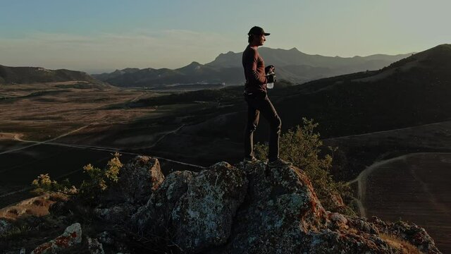 A young man in a baseball cap has reached the top of the mountain and drinks from a transparent plastic bottle on the background of a mountain landscape of the endless blue sky and sunset. Aerial view