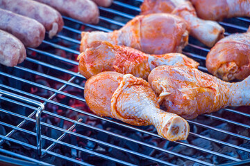Process cooking chicken drumstick and sausages on barbecue grill outdoors. Picnic, eating outdoors. Metal barbecue grill (brazier). Raw meat.