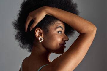 Portrait of beautiful young mixed race woman with afro hairstyle smiling, touching her hair isolated over grey background