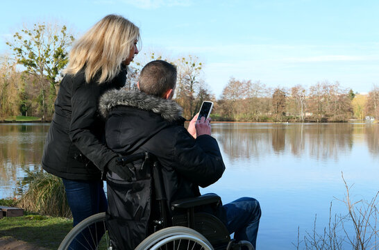 Concept of a person with a physical disability. A man in a wheelchair with a woman standing beside him. Couple using technology while looking at a smartphone. Rural scene by a lake.