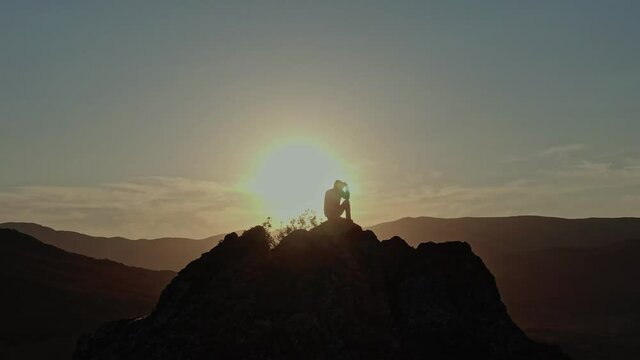 A Man Sits On Top Of A Mountain Which Is Illuminated By The Last Rays Of The Sunset. Silhouette of the Traveler on the Background of the Sun Drive. Aerial view. Surveying by drone with flying in a cir