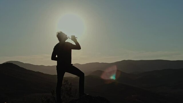 Young Man Reached the Top of the Mountain and Drinks From a Transparent Plastic Bottle Against the Background of a Mountain Landscape and Endless Blue Sky. Aerial view. Surveying by drone with flying 