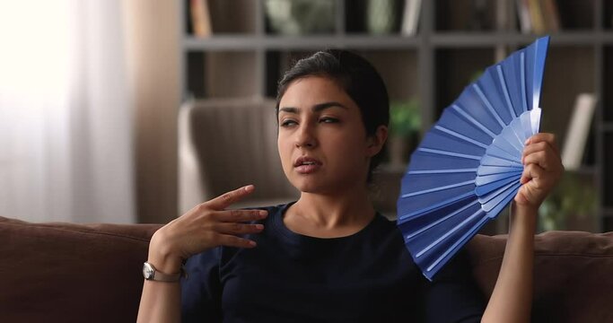 Overheated unhappy frustrated young indian ethnicity woman blowing fresh air with paper fan, sitting on sofa in living room, suffering from high temperature indoors, feeling exhausted alone at home.