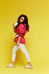Fototapeta na wymiar Full-length studio shot of the stylish model woman in pink hoodie and white pants holding a paper coffee cup and enjoying the beverage, isolated on yellow background