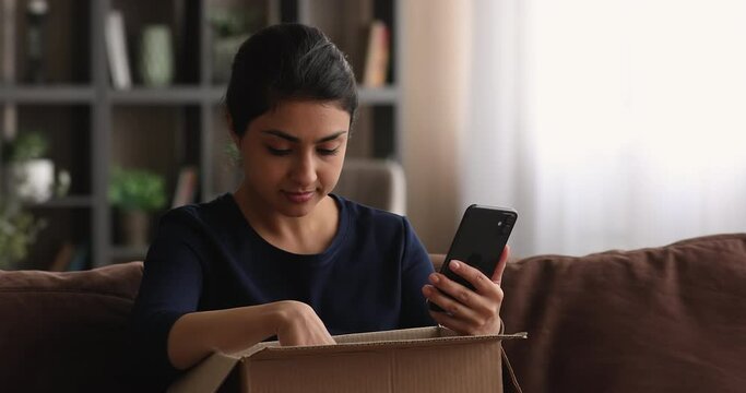 Smiling millennial mixed race indian woman unwrapping cardboard parcel at home, checking order in mobile shopping application, writing positive review, satisfied with fast courier delivery service.