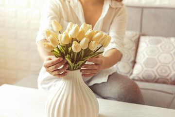 Woman putting white tulips flowers in vase sitting at the living room coffee table. Composing bouquet. Lifestyle