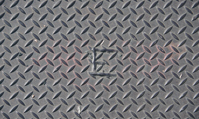 High angle full frame close-up view of a steel plate cover on a sidewalk with the letter E welded on
