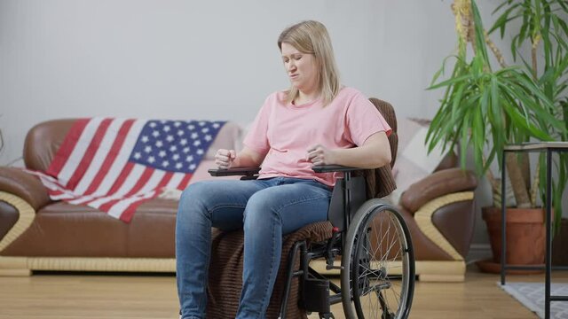 Wide shot of angry frustrated disabled American woman hitting wheelchair with hands. Portrait of depressed stressed paraplegic invalid in living room at home. Disability and anger concept.