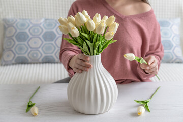 Woman putting white tulips flowers in vase sitting at the living room coffee table. Composing bouquet. Lifestyle