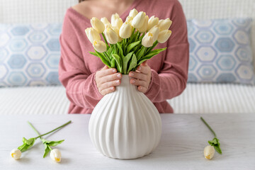 Woman putting white tulips flowers in vase sitting at the living room coffee table. Composing bouquet. Lifestyle