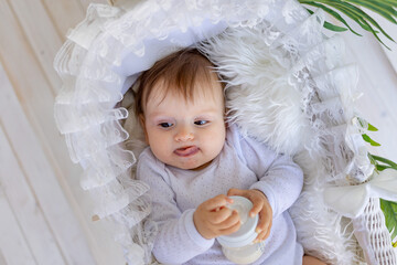 a small baby girl lies in a beautiful cradle in a white bodysuit at home and holds a bottle of milk...