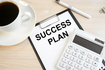 Next to a white cup of coffee and a white calculator with a pen on a wooden table is a stationery tablet on which the inscription success plan.