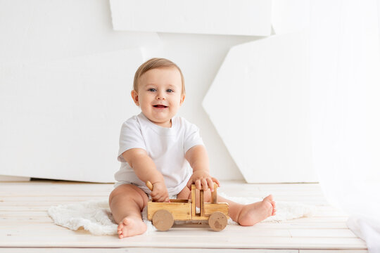 happy cute little baby six months old in a white t-shirt and diapers sits on a light background at home and plays with a wooden typewriter, space for text