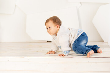 Naklejka premium a small child six months old in a knitted warm jacket and blue jeans crawls at home in a bright room