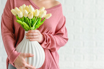 Woman in a pink pullover holding vase with white tulips flowers opposite brick wall. Copy space