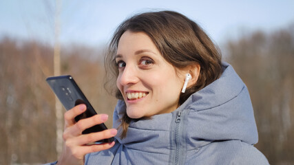 Young woman with wireless headphone is using her smartphone to send a voice message to a new social network Clubhouse standing somewhere outdoors at the park