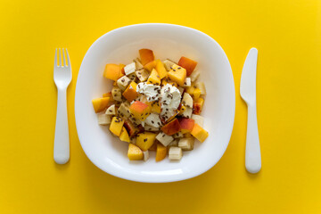 Bowl of healthy salad from fresh fruit on yellow. Healthy nutrition fruit salad with soft cheese and flax seeds in white plate on yellow background with white knife and fork.Healthy organic food.