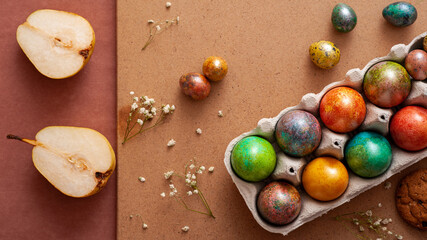 Happy Easter 2021. Colored painted eggs in a wicker basket on a brown background. Easter eggs and sweets. Cookies, pears, and chocolate.