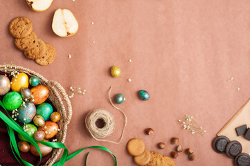 Fototapeta na wymiar Happy Easter 2021. Colored painted eggs in a wicker basket on a brown background. Easter eggs and sweets. Cookies, pears, and chocolate.