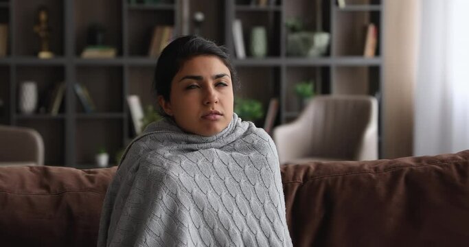Unhappy young indian ethnicity woman shivering sitting on sofa covered in warm knitted plaid, suffering from cold winter weather or low temperature indoors without heating system, feeling stressed.