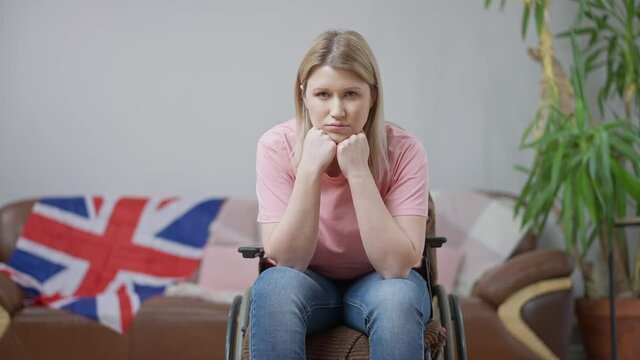 Portrait of sad young paralyzed woman in wheelchair looking at camera with UK flag at background on couch. Upset British paraplegic invalid alone at home. Disability lifestyle.