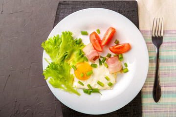 Scrambled eggs with bacon garnished with tomato and green onions and salad on a gray concrete background on a wooden stand next to a fork.