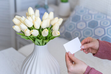 Woman pulling blank greeting card from bouquet of white tulips flowers. Valentines day. Copy space. Mock up