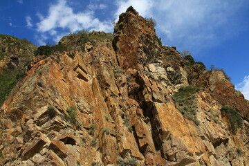 Bottom view of a high yellow-brown steep rock with an uneven stepped surface against the background of a sky with clouds on the road to the Assy plateau, bunches of grass grow on the rock,summer,sunny