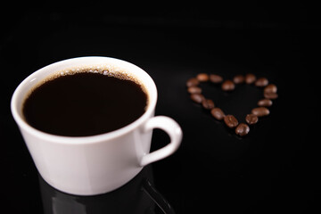 Roasted coffee beans in heart shape and white mug on black background