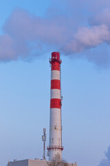 red and white chimney of a factory, from which gray smoke comes out against a blue sky on a sunny frosty winter day