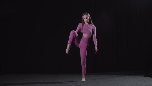 Young beautiful blonde female gymnast walks in the black studio and stretching her leg up to her head