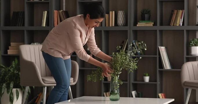 Smiling tidy young indian ethnicity female homeowner putting vase with greens and flowers on table in modern living room, making interior more comfortable, decorating styling improving accommodation.