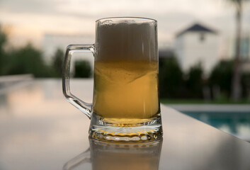 Summer vibes. Closeup view of cold beer mug on the table. The foam, golden liquid and glass on the table in the garden at sunset.