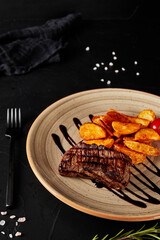grilled meat with rustic potatoes with sauce in a brown plate on a black textured background