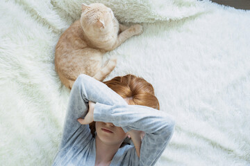 Red-haired young girl covering eyes with hands, lying on plaid with cat, suffer from insomnia headache or migraine