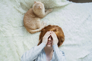 Lazy young red-haired girl wallowing on bed with fluffy ginger cat suffer from headache, hangover cover face with hands