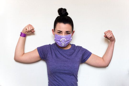 Mujer joven levantando los brazos haciendo el gesto de fuerza. Lleva camiseta morada y una mascarilla desechable con el s&iacute;mbolo de la mujer. D&iacute;a Internacional de la Mujer, 8 Marzo. Concepto feminismo.