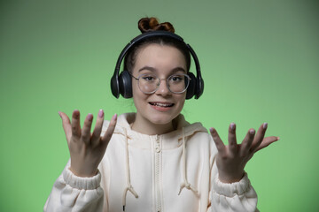Bewildered young teen girl in headphones and glasses spread hands on green studio background with copy space for ad