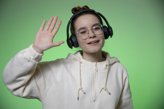 Smiling young teen girl blogger in glasses and modern wireless headphones on green studio background greeting wave hand