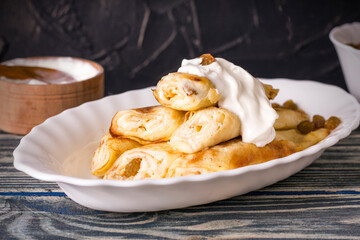 Rolled up thin pancakes stuffed with cottage cheese with raisins and sour cream on wooden table.