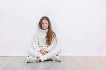 white girl 10 years old in a white sweater, jeans and sneakers sits on the floor against a white wall