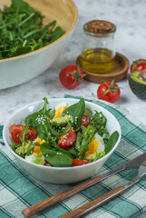 fresh greens salad - rucolla and spinach, with egg and avocado, garnished with tomato and sesame seeds in a white bowl on a light background and a bright napkin with cutlery and butter