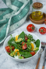 fresh greens salad - rucolla and spinach, with egg and avocado, garnished with tomato and sesame seeds in a white bowl on a light background and a bright napkin with cutlery and butter