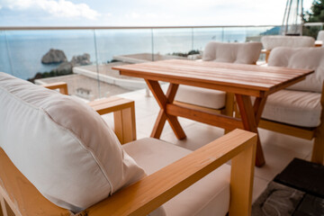 Restaurant with sea view. You can see rocks in the sea. Wooden tables and chairs with white cushions. Overcast, cloudy weather.