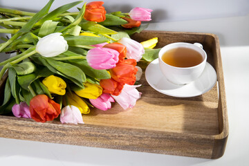 A bouquet of multicolored tulips and a cup of tea are on a tray on the table