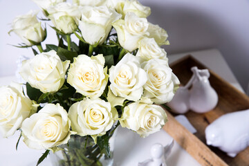A bouquet of white roses in a vintage glass vase, a wooden tray with a cup of tea, a faience figurine of an angel. Greeting card for mother's day.