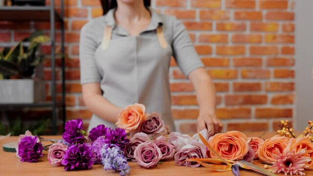 Woman professional take flowers and makes bouquet. Slow motion. Close up young female florist in apron at workplace in flower shop. Brunette girl making small bunch of flowers in flower store.