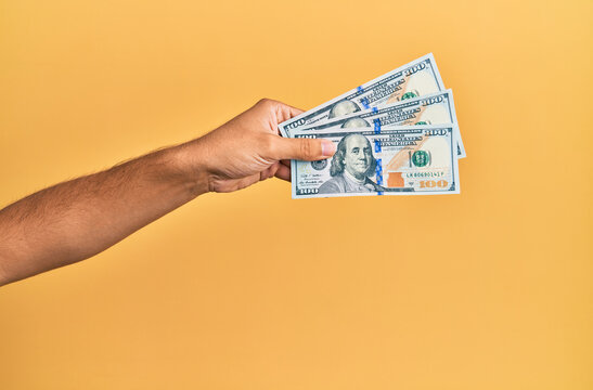 Hand of hispanic man holding 100 dollars banknotes over isolated yellow background.