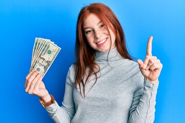 Young red head girl holding 20 dollars banknotes smiling with an idea or question pointing finger with happy face, number one